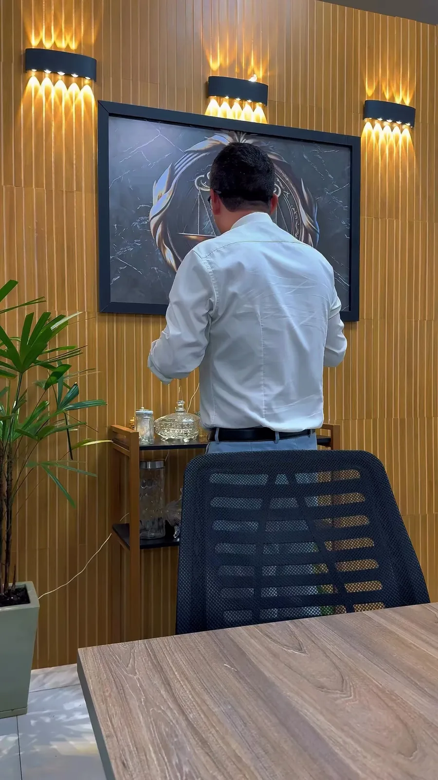 A sala do escritório: parede de madeira ripada com três arandelas acesas, um vaso de planta, a cadeira e a mesa; o Dr. Gilbran, de costas, serve o café no aparador