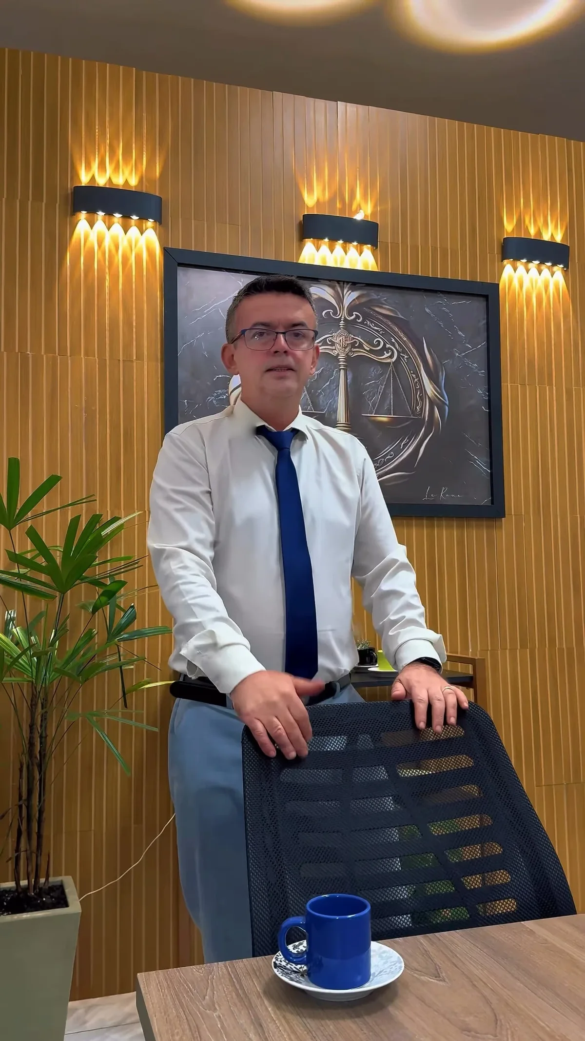 Dr. Gilbran Federici Almeida em pé no escritório, de camisa branca e gravata azul, diante da parede de madeira ripada com arandelas acesas