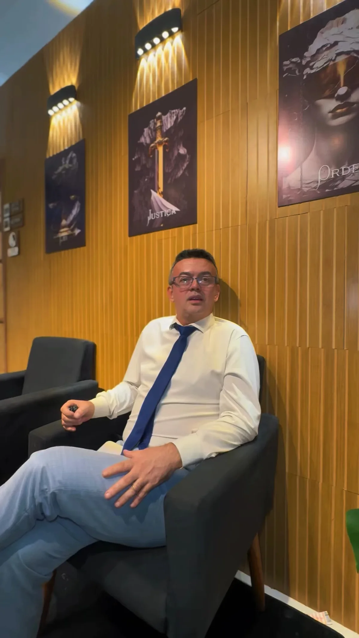 Dr. Gilbran Federici Almeida sentado numa poltrona escura do escritório, de camisa branca e gravata azul, escutando alguém fora do quadro, diante da parede de madeira ripada